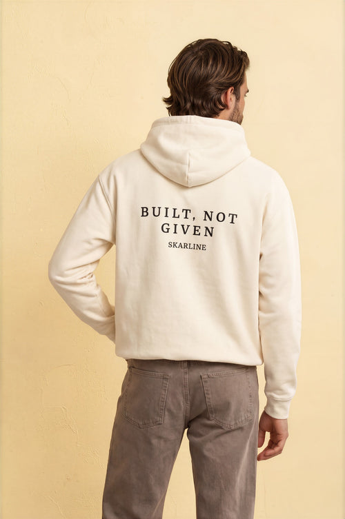 Person wearing a white hoodie with 'Built, Not Given' text on the back against a beige background